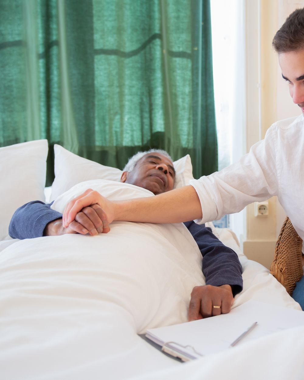A carer sits close beside an older man resting in bed, holding his hand.