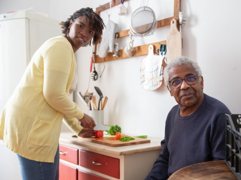 A carer prepares vegetables in a home kitchen while an older man sits nearby.
