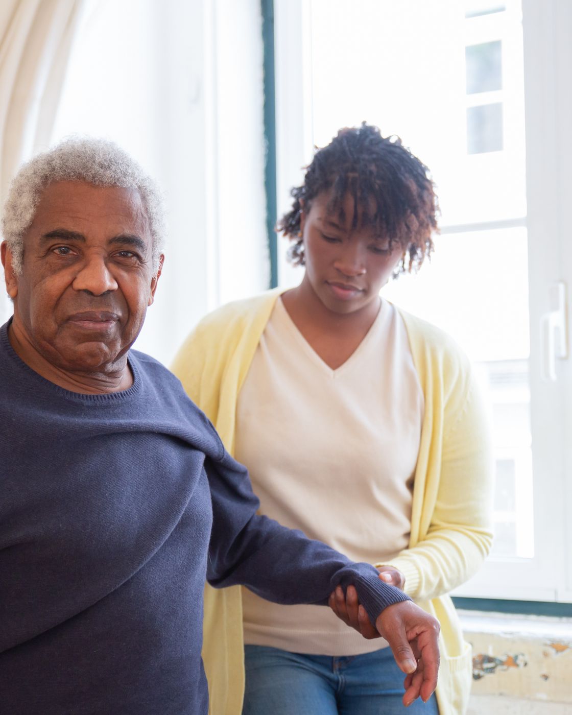 A carer stands beside an older man at a window in his home, gently holding his arm as he looks outside.