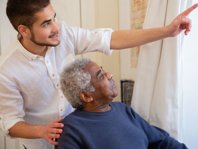 A carer gently supporting an older woman's arm as they move together inside a home.