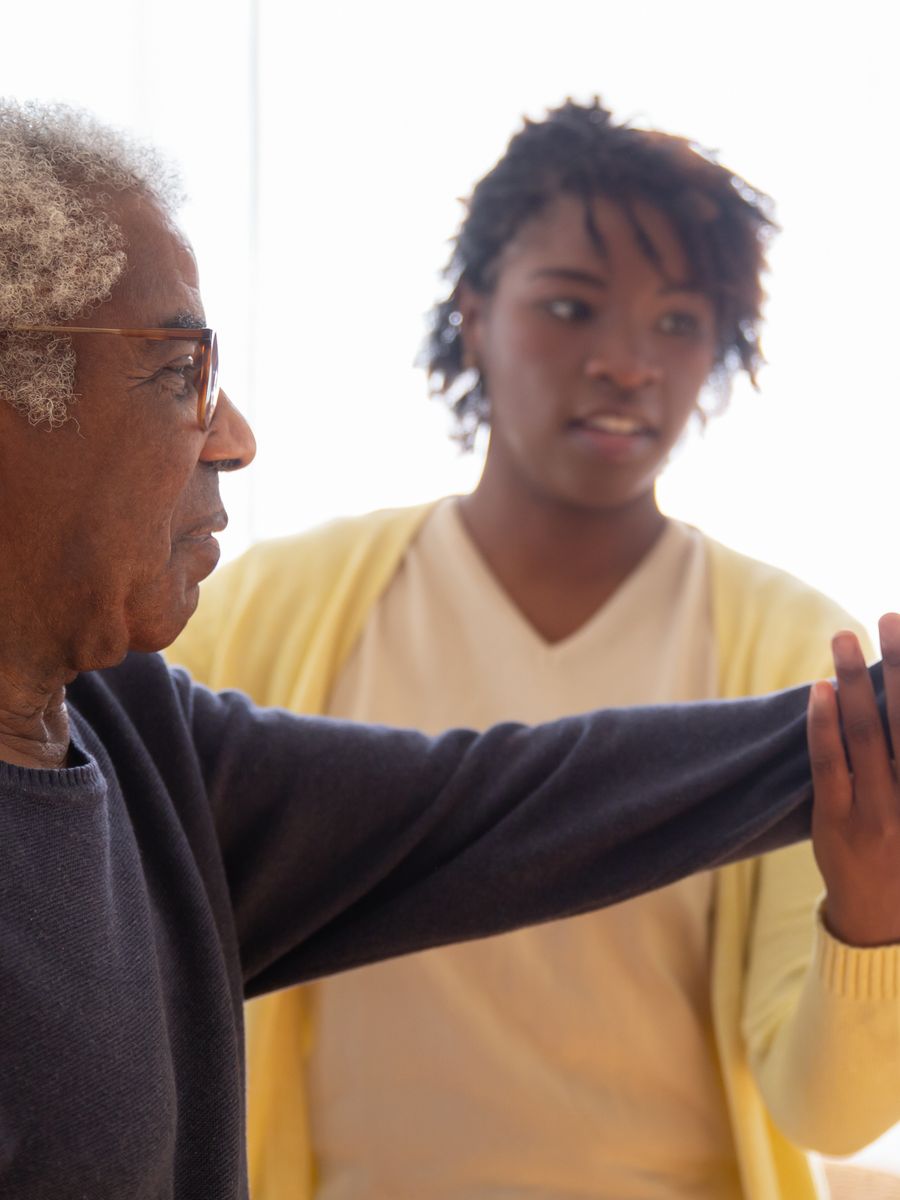 A carer helps an older man raise his arms in a gentle exercise at home.